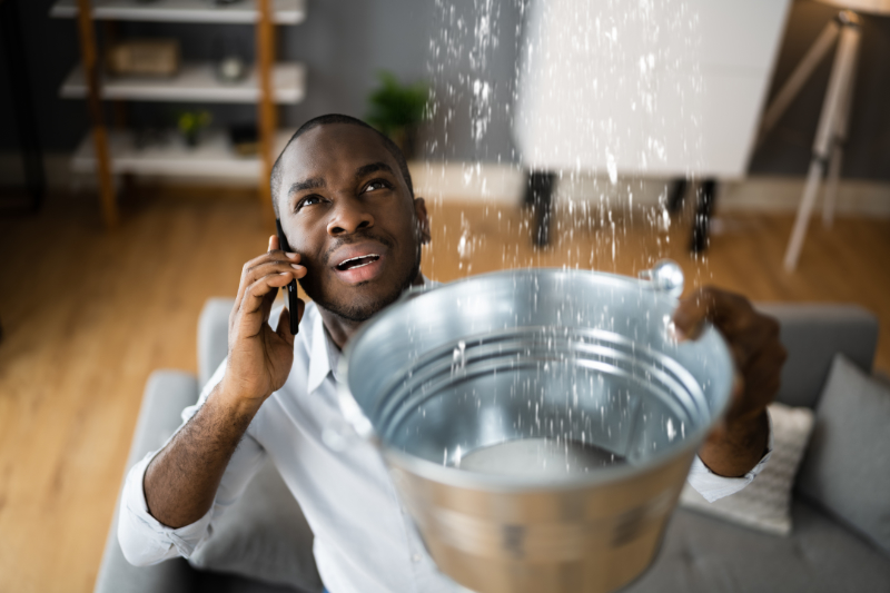 How Can I Avoid a Plumbing Emergency? A man calling a plumber while water is leaking from the ceiling into a bucket.